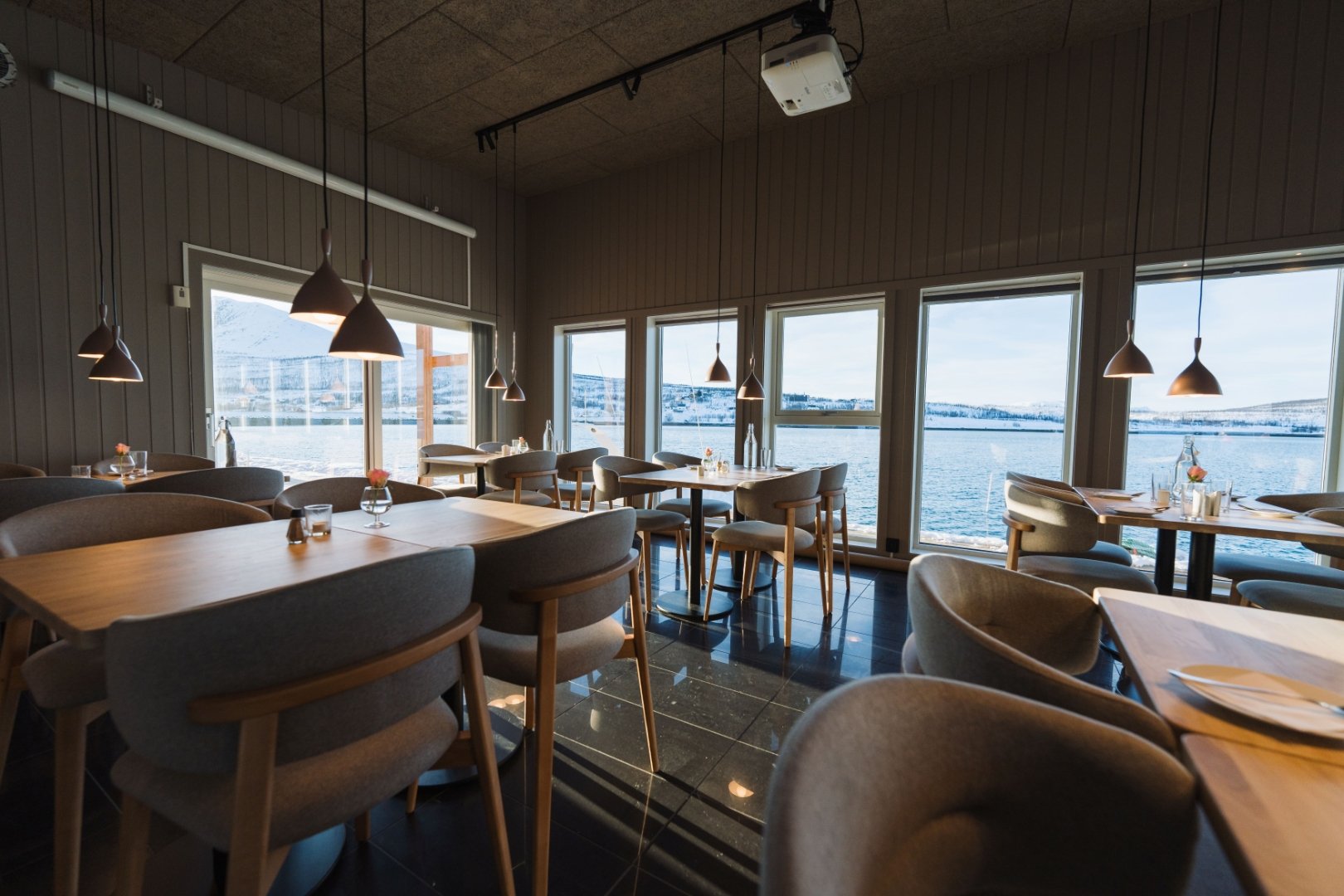 Restaurant med bord og stoler forran et panorama vindu med utsikt til sjøen. @christeradahl