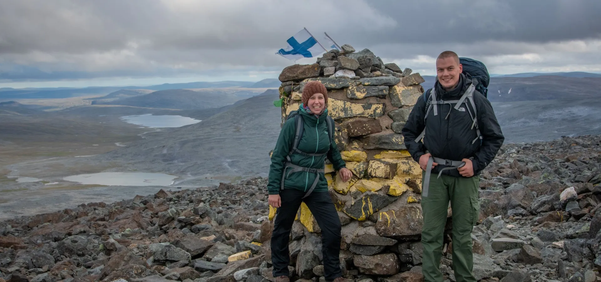 Two people standing in front of a huge rock border cairn on mountain top