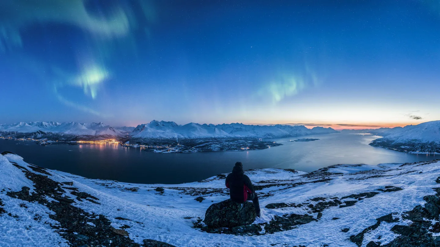 Nordlys over Lyngenfjorden, mann sitter på en fjelltopp og ser ut over fjorden